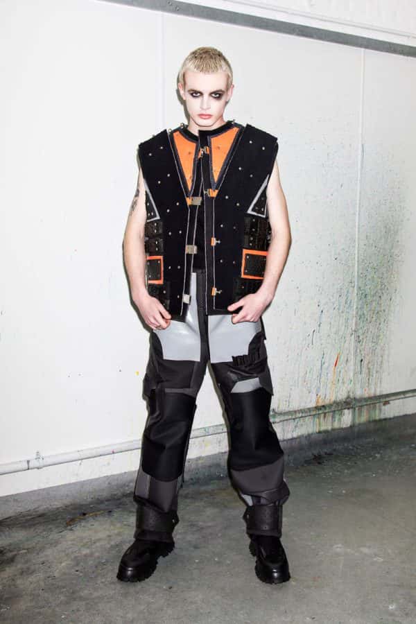 Chloe Last - White male model wears a sleeveless black and neon orange jacket, with industrial-style studded embellishment and geometric cuts of fabric. His trousers are black and grey, made from precisely cut shapes, with a variety of textures and buckles. Designed by BA Fashion student Chloe Last