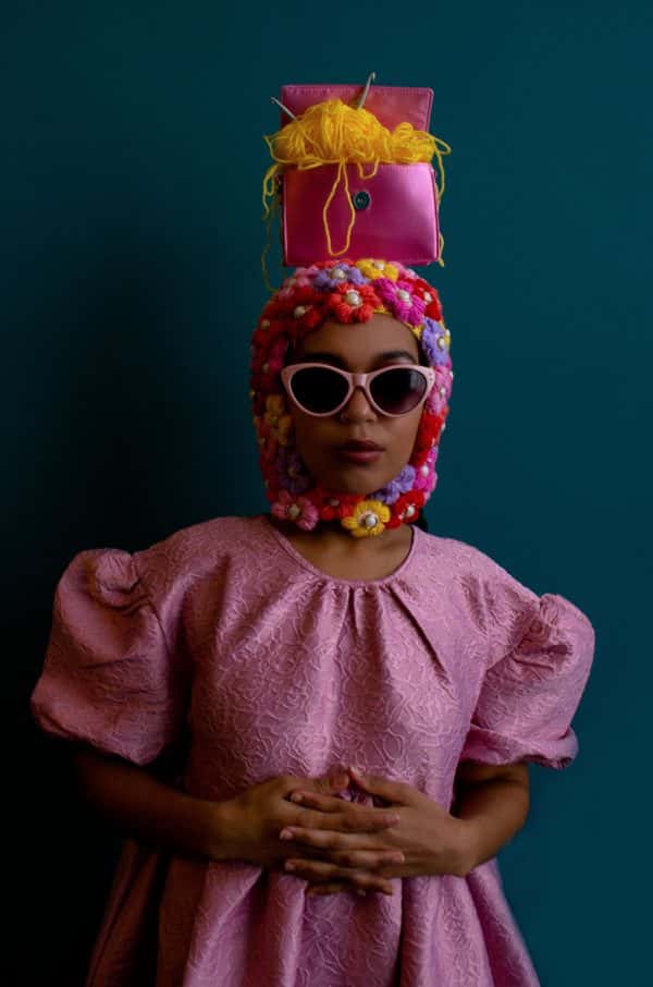 Emily Mitchell - Female model is photographed against a teal blue background, wearing a pink dress. On her head is a knitted balaclava, made out of lots of fabric flowers. On top of her head balances a small pink purse, with yellow yarn spilling out. Styled and photographed by BA Fashion Communication and Promotion student Emily Mitchell