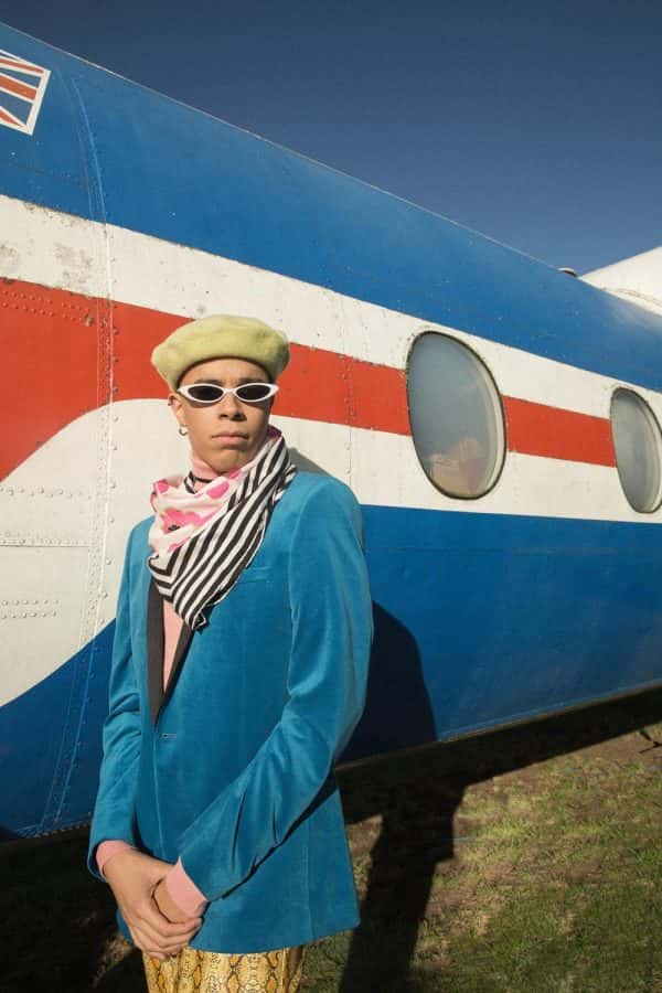 Jess Anderson - 80's style photo of a mixed race male model standing in front of a vintage aircraft. He wears a blue velvet blazer, with a patterned scarf, sunglasses and beige hat. Work by BA Fashion Communication and Promotion student Jess Anderson