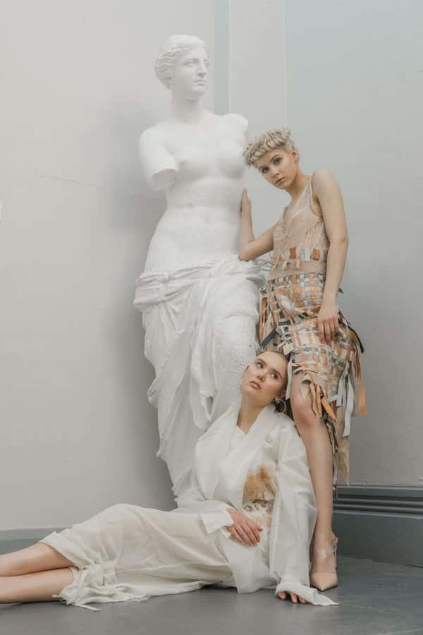 Sophie Anderson - A photograph of two female models and a statue in a grey corridor. One model is standing and leaning on the statue wearing a sleeveless textured dress, the other model is laying at her feet, wearing all white. By BA Fashion Communication and Promotion student Sophie Anderson