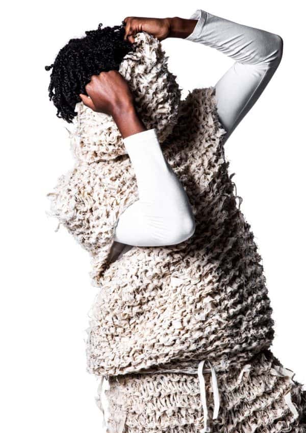 Hillary Marerwa - Black male model wearing a long sleeved white t-shirt, with a sheep's wool-like knitted short sleeved jumper over the top, with matching wool shorts. The model is pulling the loose collar up over his face. Garment designed by BA Fashion student Hillary Marerwa