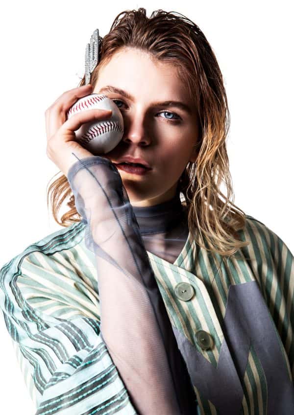 Molly Mackie - White female model is wearing a baseball style yellow/green/grey striped shirt, with a mint green M on the front, looking directly into camera. Under the shirt she is wearing a greay mesh long sleeved tshirt. She holds a baseball up, covering her right eye. Garment designed by BA Fashion student Molly Mackie