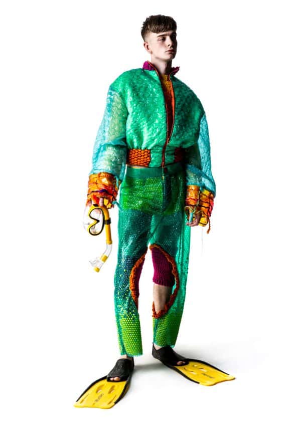 Phoebe Constable - White male model wearing a green and orange jacket and trousers with splits made from bioplastics; the trousers look similar to bubble wrap. Under the trousers are pink knitted shorts. The model wears yellow flippers for shoes and is holding goggles. Garment designed by BA Fashion student Phoebe Constable