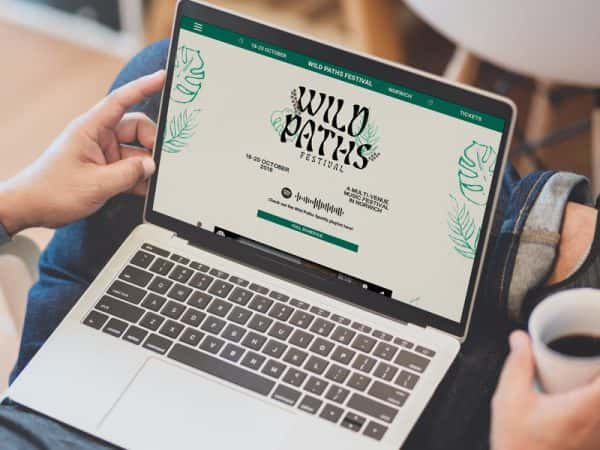 Jamie Greengrass - Website design for Wild Paths music festival in Norwich, by BA Graphic Communication student Jamie Greengrass. A person has a laptop open on a cream and green coloured website saying 'Wild Paths' with links to the full lineup.