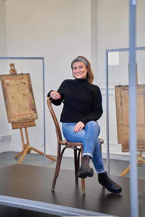 Claire allergen sitting on wooden chair with legs crossed and one arm on the back of chair on black platform. Claire has jeans and black jumper on. 2 artist easels in background.