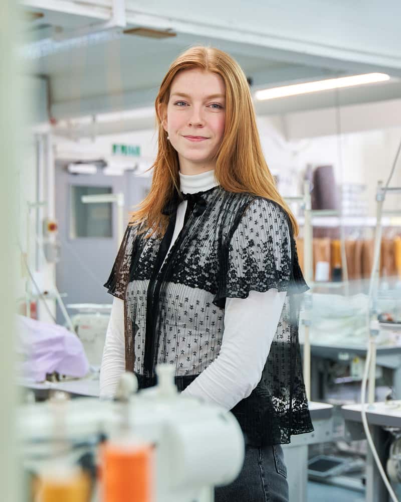 Photo of a young red haired woman standing amongst sewing machines in a fashion studio, looking into camera