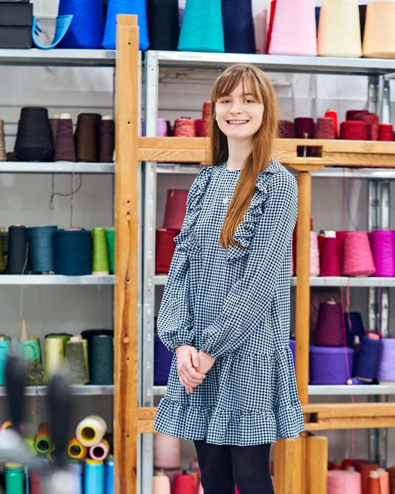 Photograph of a young woman with long hair, standing in the Textile Design workshop in front of multicoloured yarn and thread