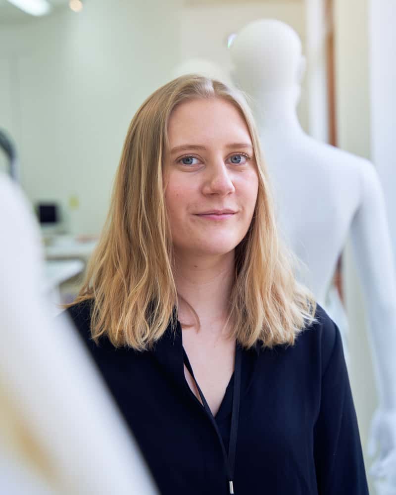A young woman with dirty-blonde hair is standing amongst mannequins in the fashion studio, looking and smiling at camera