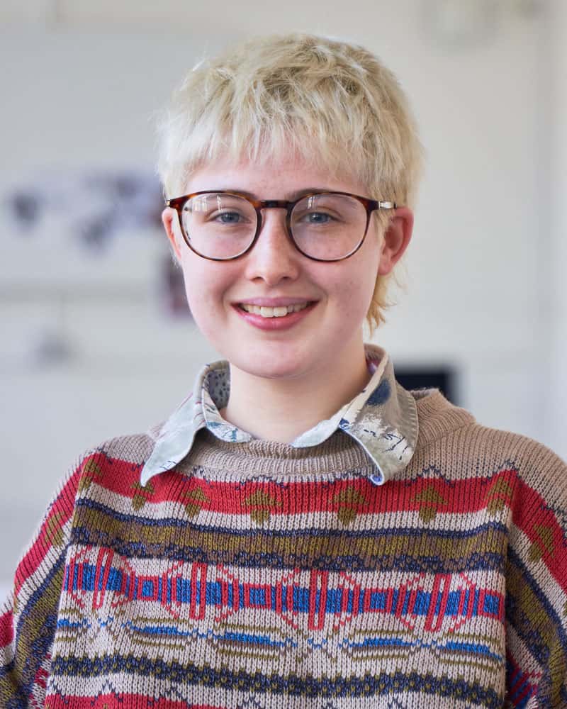 Portrait of a young person with short blonde hair and glasses looking into camera