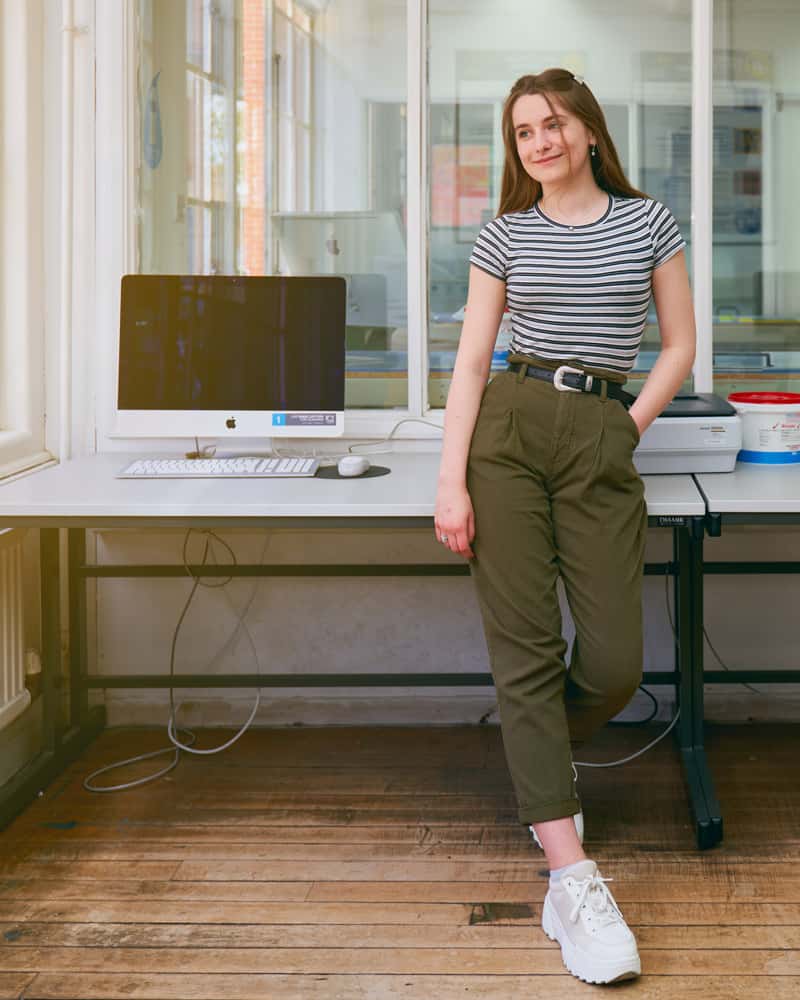 BA Design for Publishing student Sophie Ebbage in the digital design studio, perching on a desk next to an Apple iMac, looking out the window