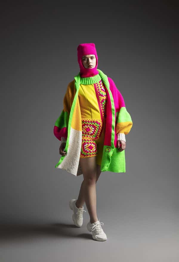 Sophie Vivien Carder - Mid-stride, the model wears a knitted jacket and open-face ski mask. The outfit is blocks of neon green, neon pink, and bright yellow; whereas the knitted shorts and top have circular checkered style patterns.