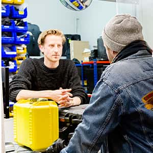A student in denim jacket and grey beanie picks up equipment from the media resource. A bright yellow box sits on the counter and in the background there are racks of hardware and accessories for students to borrow.