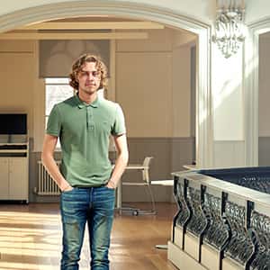 Bradley Fletcher stands in the architecture studio. Bradley has long blonde hair and wears a green polo shirt and blue jeans.