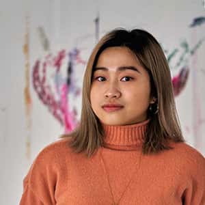 Yuyu Tse wears an orange roll neck jumper, and stands in front of a colourful pattern on the wall