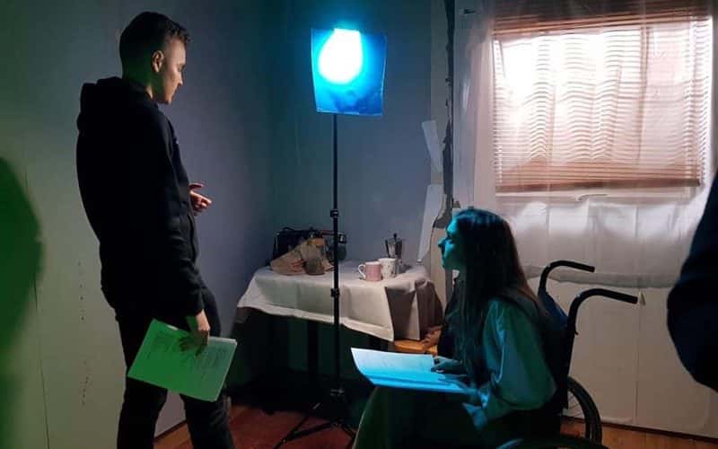 Image of two people reading scripts in a dark room in a house in a film set. The curtains are closed and there is a table with refreashments on. One person is in a wheelchair.