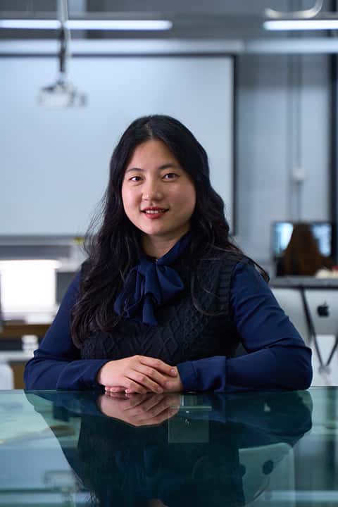 Portrait of Senior Computer Science Lecturer Wenshu Zhang in the Digital Darkroom. Wenshu has long black hair, and wears a dark navy cable knit sweater vest over a long sleeved dark blue blouse with tie neck