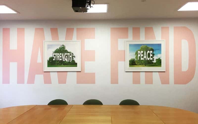 Photograph of a conference room with a large table and chairs in the centre. On the walls is large lettering, spelling out 'Have' and 'Find', coloured pale pink. In two white picture frame centred over each of the words, is a photograph of a tree, with the word 'Strength' in one frame, and the other the word 'Peace'. The work was created by Carl Rowe, for Norwich University of the Arts' research output.